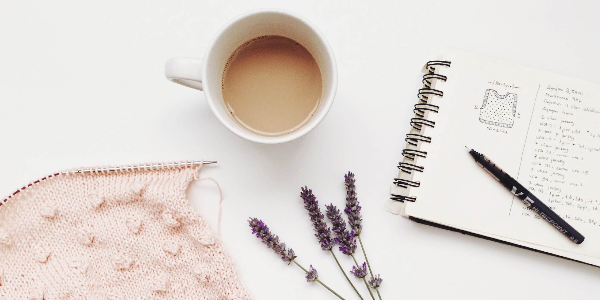 Knitting project, a notebook, cup of coffee, and dried lavender resting on a white surface