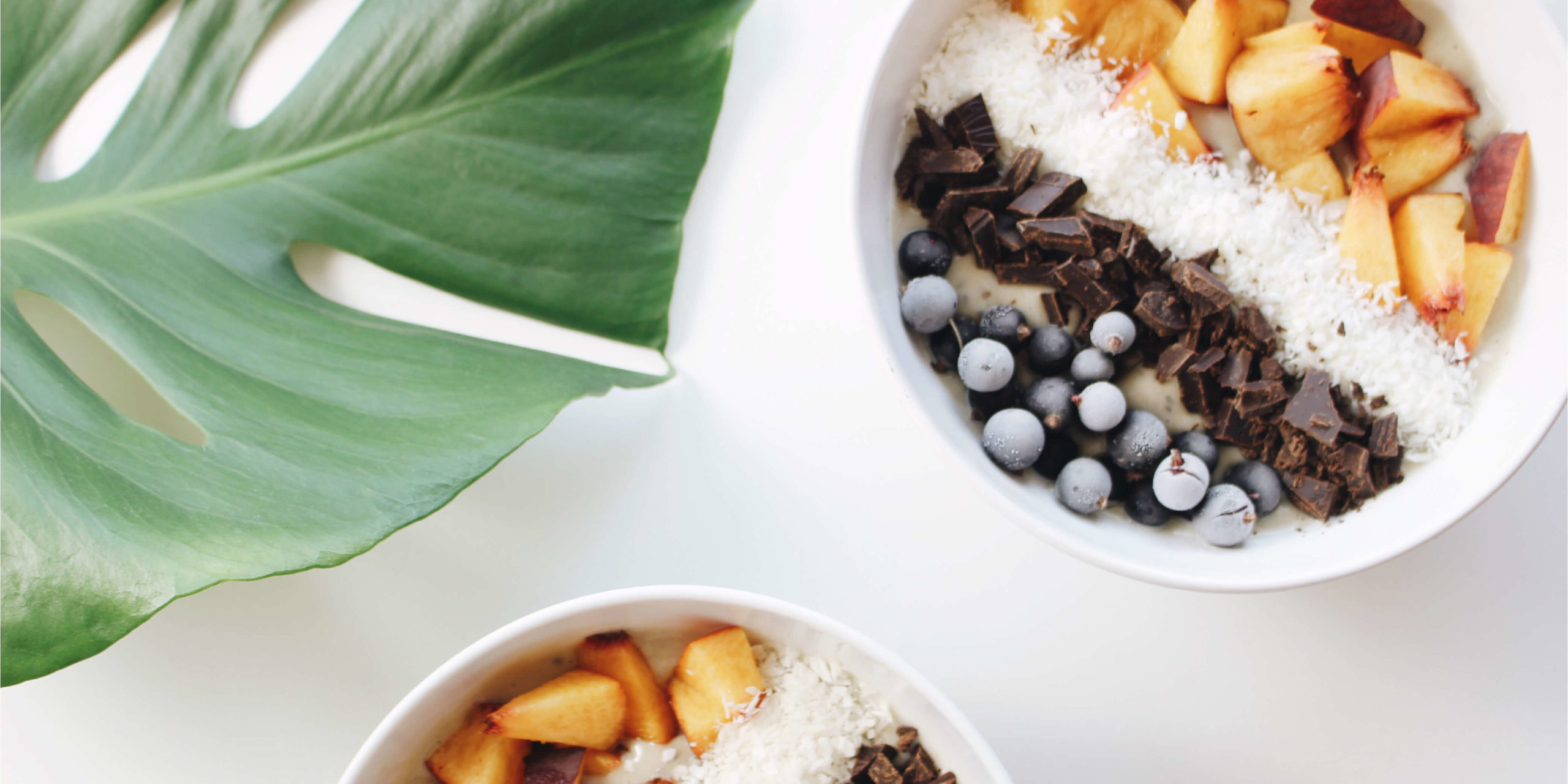 Monstera leaf beside smoothie bowls filled with frozen chopped peach, shredded coconut, dark chocolate and blackcurrants