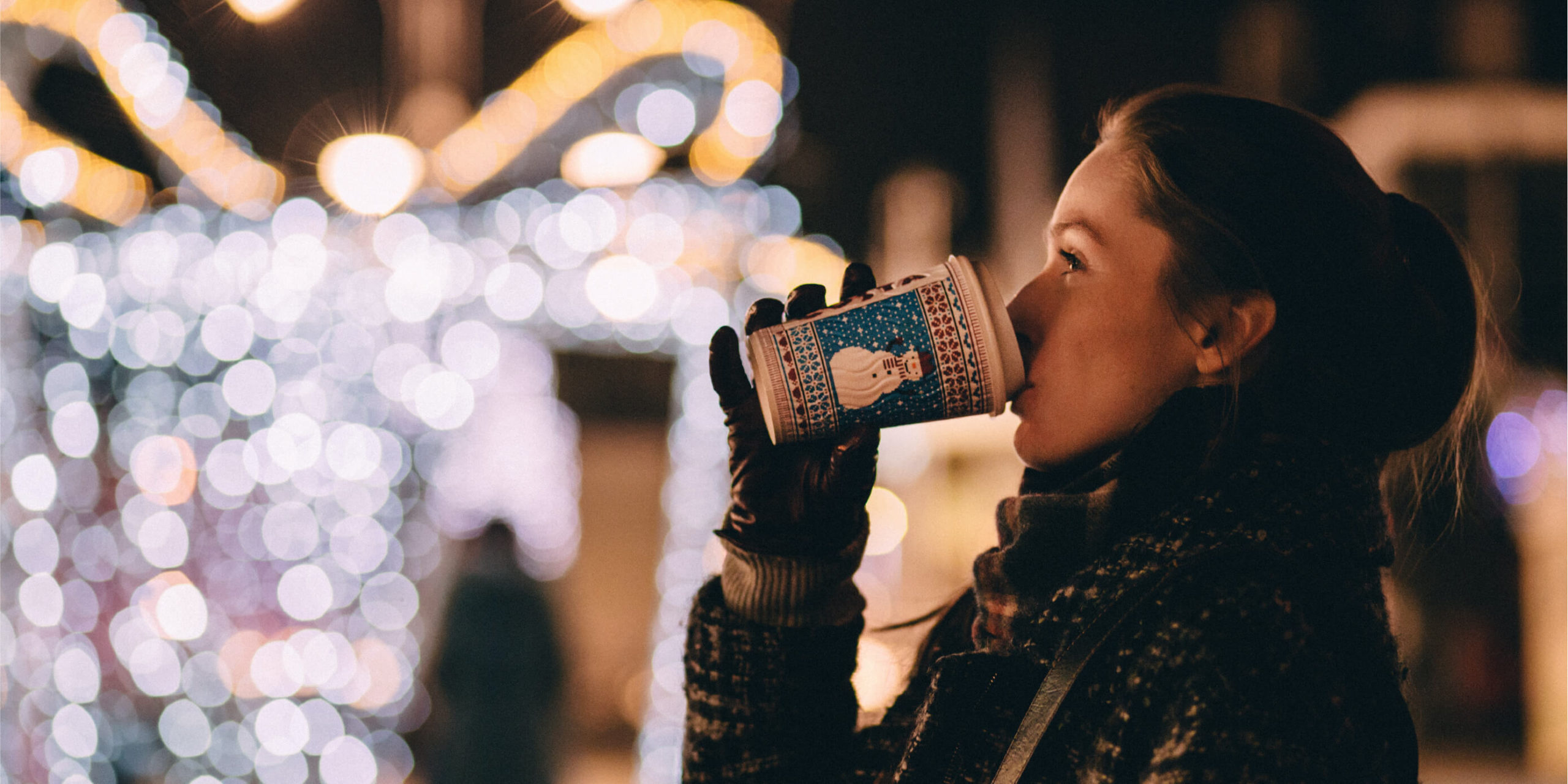Woman drinking coffee surrounded by outdoor holiday lights.